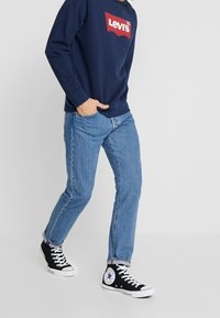 Marinblå sweatshirt med röd Levi's-logga, ljusblå jeans och svarta högklackade sneakers med vita detaljer. Avslappnad outfit för vardagsbruk.