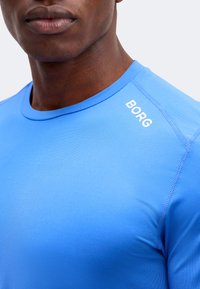 Κοντινό πλάνο ενός ατόμου που φοράει ένα μπλε αθλητικό μπλουζάκι με το λευκό «BORG» τυπωμένο κοντά στον δεξιό ώμο.