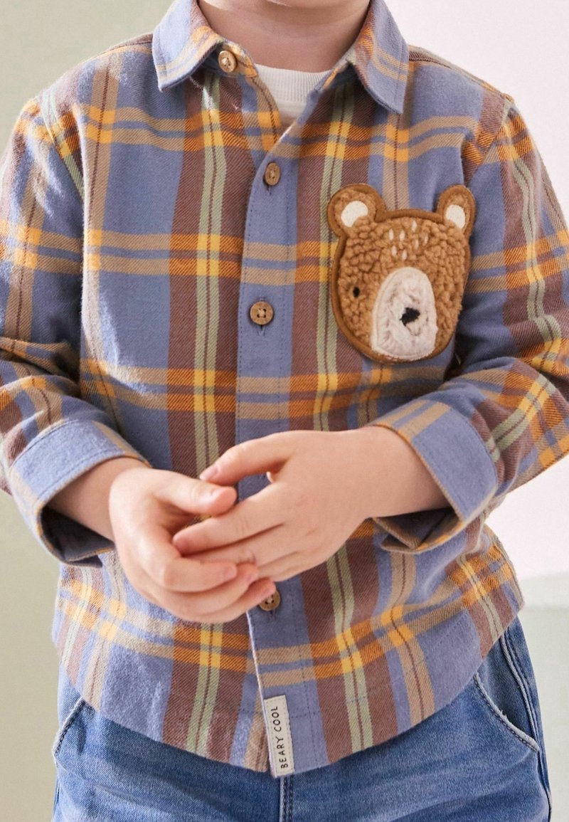 Jeune enfant portant une chemise à carreaux bleue et orange avec un écusson ours sur la poitrine et un jean bleu, les mains délicatement jointes devant.