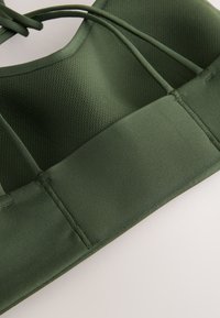 Nahaufnahme eines dunkelgrünen Sport-BHs mit Mesh-Gewebe am oberen Teil und glattem Material am unteren Bund mit doppelten Schulterträgern.