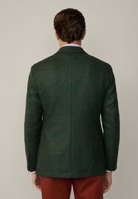 Grön tweedblazer med rutmönster, skräddarsydd passform, enkel sprund och bruna accenter; bärs över en ljusblå rutiga skjorta och röda byxor.