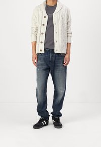 Off-white stickad cardigan med kabelmönster, grå skjorta, lösa blå denimjeans och svarta sneakers med vita ränder.