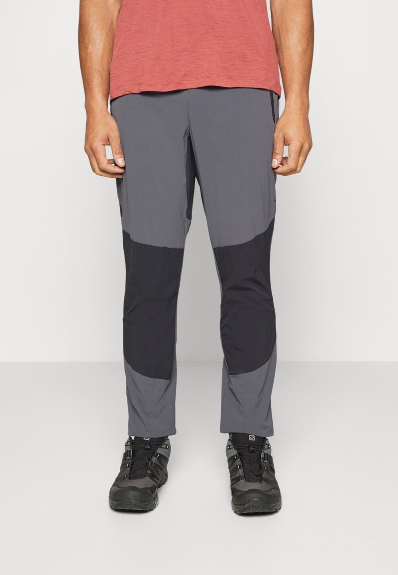 Mann trägt zweifarbige Outdoorhose in Grau und Schwarz mit schwarzen Wanderschuhen, steht vor einem schlichten weißen Hintergrund.