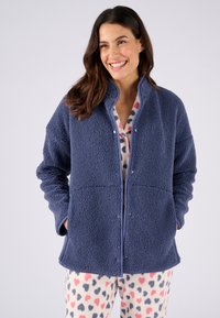 Marineblauwe fleecejack met een gestructureerd oppervlak, drukknoopsluiting, opstaande kraag en twee zijzakken, gelaagd over een geblokte pyjamabroek.