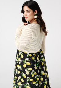 Une femme aux longs cheveux bruns porte un pull crémeux à texture sur une robe à motifs de citrons, ornée de boucles d'oreilles florales, se retournant par-dessus son épaule.