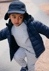 En ung dreng iført en marinebl�å puffjakke og hat, lys grå træningsdragt og marineblå sneakers, står udendørs på en legeplads.