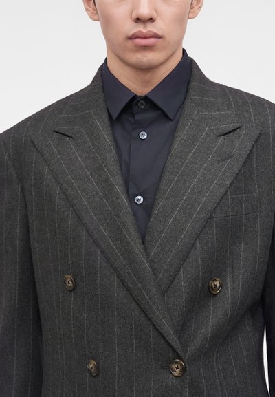 Homme portant un blazer croisé à fines rayures gris foncé sur une chemise noire boutonnée, montré du menton à la taille.