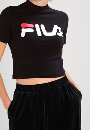 Vrouw die een nauwsluitend zwart cropped FILA T-shirt en losse zwarte fluwelen broek draagt met handen in de zakken tegen een effen achtergrond.