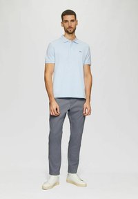 Mann steht aufrecht in einem hellblauen Poloshirt, grauen Hosen und weißen Sneakern vor einem einfarbigen hellen Hintergrund.