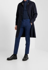 Marineblauwe wollen overjas, donker overhemd met knopen, blauw op maat gemaakte broek en zwarte leren schoenen. De jas heeft een kraag en accenten bij de voorzakken.