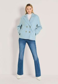 Frau in einem hellblauen doppelt-knöpfbaren Mantel, weißem Rollkragenpullover, weit geschnittenen blauen Jeans und weißen spitzen Stiefeln, die vor einem schlichten Hintergrund steht.