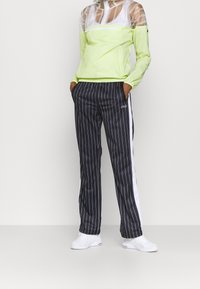 Transparentes Mesh-Oberteil mit langen Ärmeln in Neon-Grün über einer taillierten, elastisch abgesetzten Taille, kombiniert mit schwarz gestreiften Hosen mit weißen Seitenakzenten.