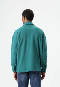 Oversized teal overhemd gemaakt van gestructureerde stof, met een relaxte pasvorm, lange mouwen en knoopmanchetten. Naden zorgen voor extra detail.