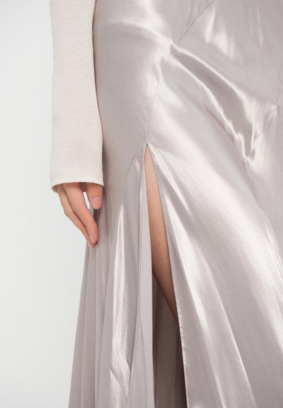 Hosszú ezüst szatén ruha combmagasságú hasítékkal, sima textúrával és finom fényes hatással, amelyet egy világos anyagból készült testhezálló hosszú ujjú felső egészít ki.