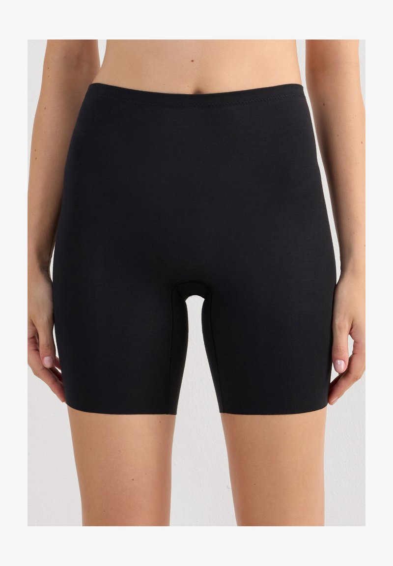 Waist to mid-thigh view van een persoon die effen zwarte, nauwsluitende shorts met een gladde tailleband draagt tegen een neutrale achtergrond.