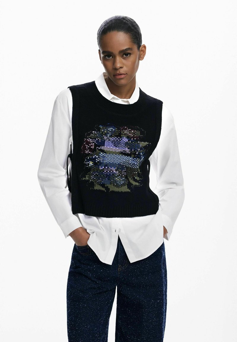 Schwarzer ärmelloser Pullover mit einem mehrfarbigen Blumenmuster, getragen über einem weißen Hemd mit Knöpfen; hat einen gerippten Halsauschnitt und seitliche Bindebänder.