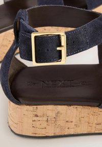 Sandale din suede de culoare bleumarin, cu toc din plută, dotate cu o cataramă din metal aurit și o talpă interioară netedă de culoare maro, inscripționată cu "NEXT."