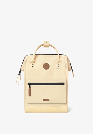 Beige canvas backpack with a top handle, black zipper accents, front zip pocket, circular logo, and textured bottom panel.