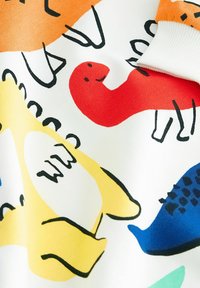 Valkoinen kangas, jossa on värikkäitä dinosauruksen kuvastoja oranssina, punaisena, keltaisena ja sinisenä. Mustat ääriviivat korostavat leikkisää muotoilua. Pehmeä rakenne.