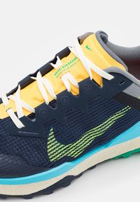 Gros plan sur une chaussure de trail multicolore avec une languette jaune, des lacets blancs, une tige en mesh bleu marine, un swoosh Nike vert et une semelle bleu-beige.