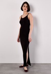 Abito lungo nero a coste con spalline sottili, spacchi laterali e silhouette aderente. La modella indossa ballerine bordeaux. Design semplice e minimalista.