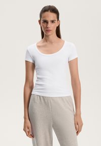 Bílý krátký rukáv s žebrováním a výstřihem do lodi, přiléhavý design a hladká textura, v kombinaci s světle šedými bavlněnými domácími kalhotami.