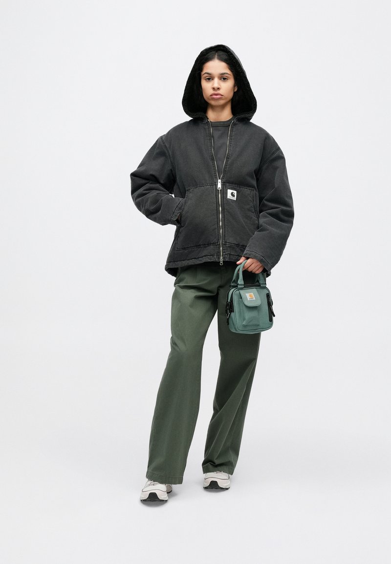 Zwarte capuchonjas met ritssluiting aan de voorkant, gecombineerd met groene wijdvallende broek en een kleine groene tas. Model staat op een effen achtergrond.