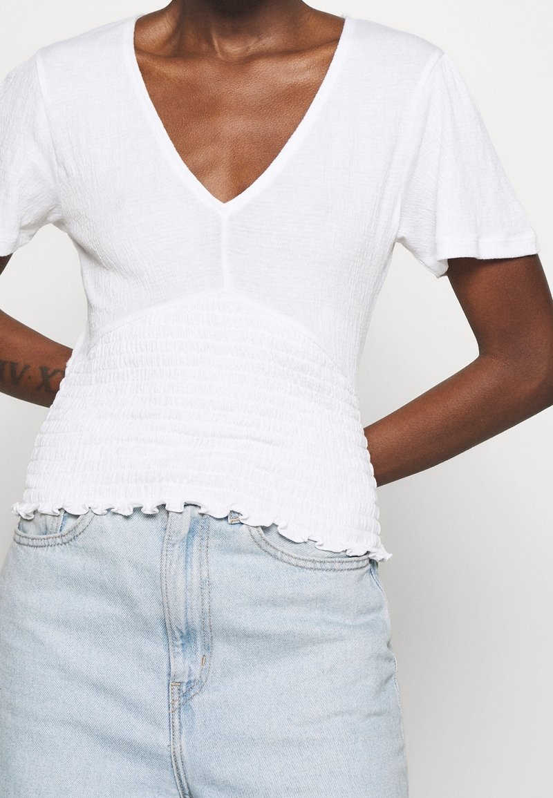 Top blanco de manga corta con escote en V y diseño fruncido texturizado en la cintura. El detalle del dobladillo deshilachado añade un toque casual. Combinado con denim claro.