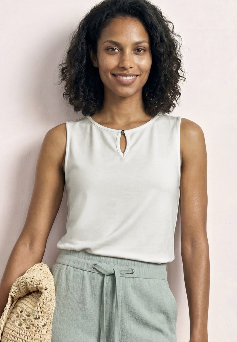 Femme aux cheveux bouclés et foncés sourit, portant un haut sans manches blanc et un pantalon vert clair, tenant un sac beige en crochet contre un mur pâle.