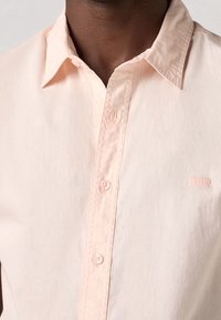 Camisa de algodón rosa claro con botones, que presenta un cuello clásico, un bolsillo en el pecho y un sutil bordado de la marca.