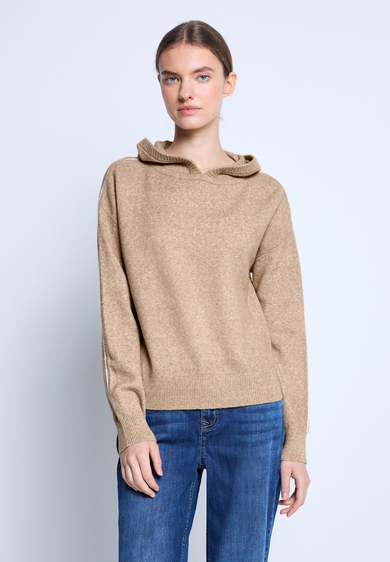 Beige gestrickter Pullover mit Kapuze, der eine strukturierte Oberfläche und einen gerippten Saum hat. Kombiniert mit blauen Jeans und einem neutralen Hintergrund.