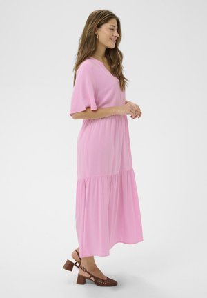 Mosolygó nő oldalról állva, világos rózsaszín, rövid ujjú midi ruhát viselve és barna, alacsony sarkú pántos cipőt hordva.