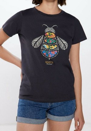 Vrouw draagt een zwart t-shirt met kleurrijk bloemig bijenontwerp en tekst "ROOTS Forever", gecombineerd met opgerolde denim shorts.