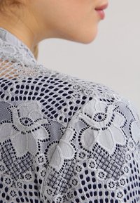 Ljusgrå spets tyg med blommiga mönster och öppna vävdetaljer, som överlagras på en mörkare marin bakgrund, visad från en axelvinkel.