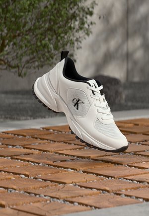 Weiße und schwarze Calvin Klein Sneaker schweben über braunen quadratischen Fliesen im Freien mit grüner Vegetation und grauer Wand im Hintergrund.