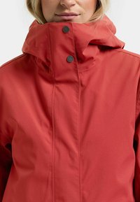 Kvinde iført en rød jakke med høj krave og to sorte trykknapper lukket ved halsen.