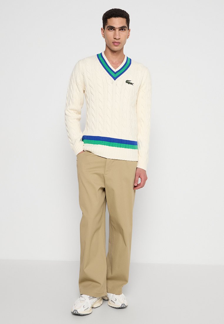 Maglione cremoso a tricot con scollo a V, con dettagli a righe blu e verdi all'orlo, abbinato a pantaloni beige e sneaker bianche.