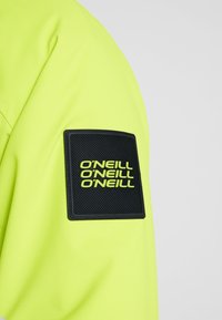 Manche de veste jaune vif avec un patch carré noir comportant le texte répété en jaune néon « O’NEILL » en lettres grasses.