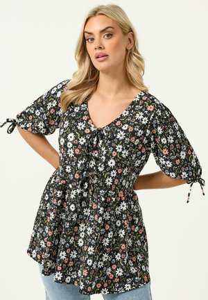 Blondynka w czarnej bluzce w kwiaty z białymi i różowymi kwiatami, z bufkami związanymi przy łokciach, stojąca z rękami na biodrach.