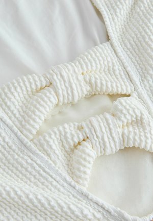 Hvid badedragt med struktureret, ribstrikket stof med markante sløjfer ved halsudskæringen. Glat inderfor i en lysere nuance.