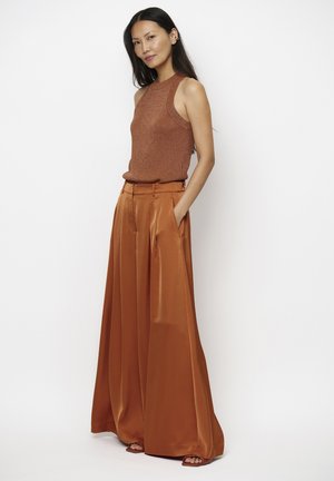 Slim woman with long black hair wearing a sleeveless rust knit top and wide-leg rust satin pants with hands in pockets.