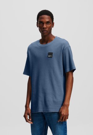 Tricou scurt cu textură în formă de vafe albastre, cu guler rotund și un patch logo negru pe piept. Asortat cu blugi albaștri.