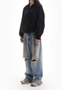 Personne portant un hoodie noir zippé, un jean large déchiré bleu clair, des baskets argentées et une bague argentée à la main gauche.