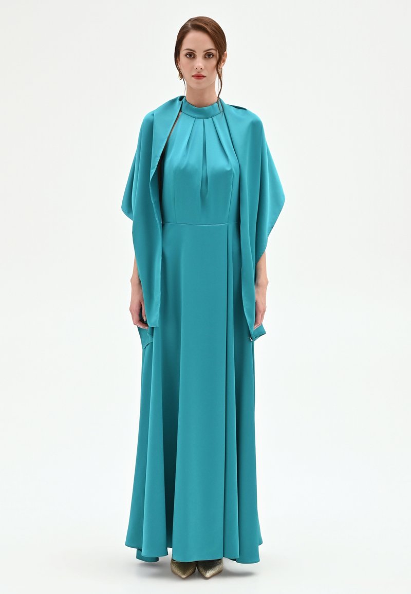 Femme portant une robe longue couleur sarcelle avec un col haut plissé et un châle ample assorti, debout devant un fond blanc uni.