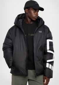 Črna puffer jakna s kapuco, s sijočim zaključkom, dvema stranskima žepoma in belim geometrijskim vzorcem na rokavu. Obuta je čez črn sweatshirt.