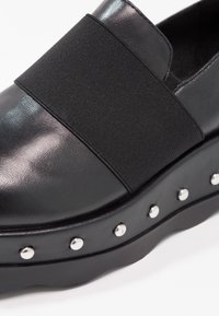Chaussure noire à enfiler avec une tige en cuir lisse et un bandeau élastique. La semelle épaisse est ornée de clous argentés sur le côté.