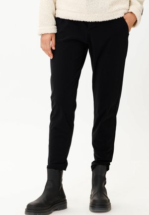 Personne portant un pantalon slim noir, des bottines noires et une veste polaire crème, une main dans la poche.