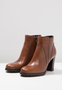 Botas de cuero marrón hasta el tobillo con un diseño elegante, cremallera lateral y una suela texturizada. El tacón es ancho y mide aproximadamente tres pulgadas de altura.