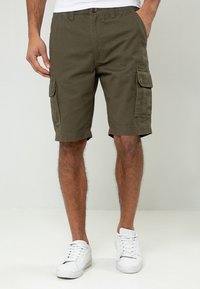 Shorts cargo verts olive en coton avec poches latérales, passants de ceinture et ourlet cousu, associés à des baskets blanches.
