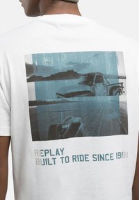 Camiseta de algodón blanca con un estampado gráfico azul de un coche y un camión vintage, con el texto "REPLAY BUILT TO RIDE SINCE 1981" debajo.
